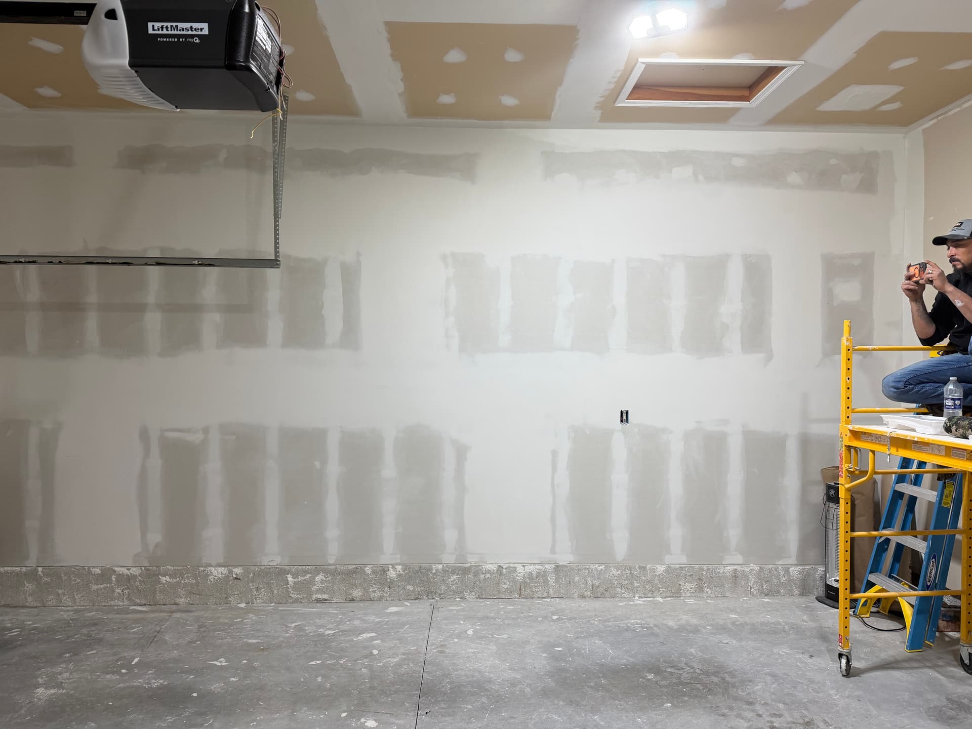 Wide garage wall fully drywalled and taped, with crew member on scaffolding